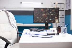 Modern clinic office sits empty, with desktop monitor displaying viral cell for biology research. Unoccupied doctors desk and technology reflect ongoing efforts in medical science against sickness. photo