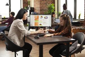 Playful daughter interrupting her mom as she tackles complex financial data on computer monitor. Caucasian little girl sitting and playing around on table, disturbing tired mother at work. photo
