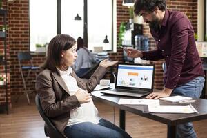 Pregnant businesswoman with laptop at desk is reviewing financial charts with male coworker. Caucasian man and expectant mother with paperwork on table, having project discussion in brick wall office. photo