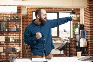Caucasian male manager dancing with excitement in startup office after securing business deal. Young man with beard punching the air with joy after successfully completing project presentation. photo