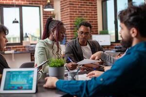 Diverse coworkers collaborate on project in modern workspace with documents and devices. Team members seated at table, discussing company tasks and reports as tablet displays data graphs in office. photo