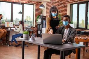 Female manager stands and points to device screen, showing male coworker the data errors in startup office. White woman with clipboard gestures to laptop as colleague listens, both wearing face mask. photo