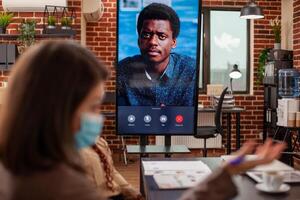 Black man seen on led screen panel, conducts videoconference call with team in brick wall office. Digital monitor shows ongoing virtual meeting of client, representing remote communication in office. photo
