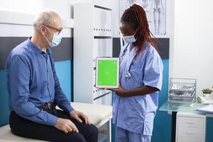 Black nurse and elderly white man looking at digital device displaying greenscreen. Healthcare specialist discussing medical results with pensioner patient using tablet with isolated copyspace. photo