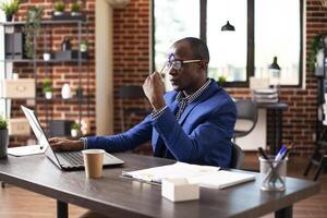 Businessman seated at table, deep in thought with pen in hand, focusing on startup research. Manager with laptop in brick wall office, tapping his forehead and studying company business analytics. photo