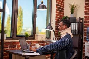 Male manager wearing cervical collar, works diligently on company research and typing on digital device at table. Young man with neck brace using laptop for reviewing business project at office desk. photo