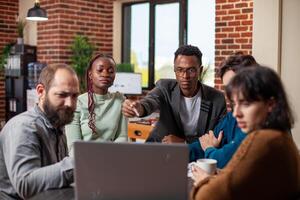 Multiethnic team gathers around laptop for discussion, showcasing focus and teamwork in modern workspace. Startup employees collaborating on project with concentration at shared device on office desk. photo