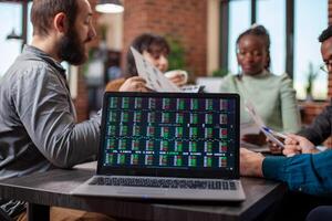 Startup team discusses documents in office as financial statistics are displayed on a laptop. Closeup on digital device on desk showing key performance indicators in brick wall workspace. photo