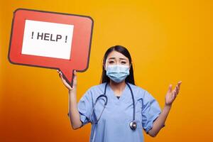 Asian female nurse wearing scrubs and a face mask, holding a help speech bubble, looking distressed. Young woman in medical uniform, grasps a red signboard with an urgent message. photo