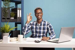 Company advisor with glasses, making an okay gesture with his fingers, looking happy and satisfied with startup project. Black male freelancer giving an approval gesture and smiling at camera. photo
