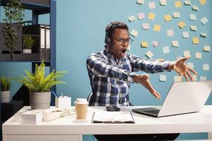 Black businessman aggressively points to device screen, reacting with rage during a heated phone call with his employees. Furious finance analyst shouting at coworkers in a meeting on his laptop. photo