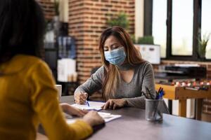 Female business advisor reading document on table and discussing strategy ideas with coworker in modern workspace. Asian recruiter interviewing a woman, asking questions about cv resume on clipboard. photo