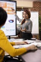 Marketing consultant standing and presenting growth data to coworker seated at office desk. Female business advisor gesturing to device screen, showing her colleague the company analytics summary. photo