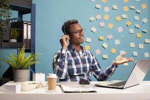 Telemarketer sitting at desk and assisting a client over the phone about application documentation. Company consultant using his laptop and handling a call, providing assistance to the employee. photo
