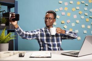 African american company advisor taking selfie photo and having a cup of tea in his modern workspace. Black businessman with cellphone is taking a of himself about to drink coffee at his desk.