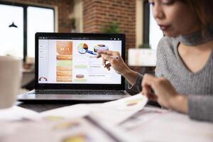 Selective focus on a digital device displaying business analytics as female analyst points at screen with a pen. Focused office worker reading her documents as she gestures towards the laptop on desk. photo