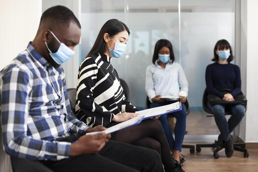 Diverse applicants with recruitment documents waiting in office lobby for interviews, preparing for hiring meetings. Multiethnic people with face masks, sitting in queue and reading their resumes. photo