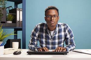 Serious male manager leaning at his office desk, typing on his computer keyboard, focused on updating company information. POV of african american analyst closely reviewing startup project results. photo