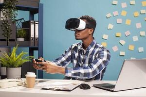 Playful male employee competing in a game using a modern console controller. Startup project manager unwinding with metaverse gaming, using a gamepad and VR headset at his office desk. photo
