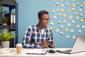Smiling male accountant holding a gamepad, enjoying a fun game on laptop, taking a break at work. Happy business advisor with a controller, having a gaming session. photo