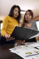 Selective focus on a clipboard being held and reviewed by female finance analysts standing by the table. Closeup of a document board in the hands of businesswomen near office desk, examining data. photo