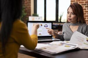Project manager seated with coworker at desk, pointing to financial graphs on laptop during their business meeting. Asian female advisor describing company analytics to employee in brick wall office. photo