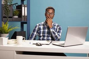 African american manager with hand on jaw, having a shocked expression, eyes wide open looking at camera. Portrait of a stunned black male employer cupping his mouth after reading a surprising email. photo