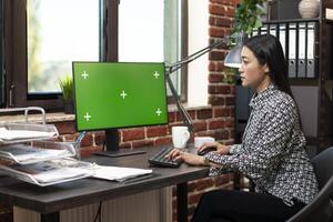 Asian female employee seated at office desk, focused on desktop monitor with a greenscreen. Businesswoman working in a brick wall workspace as computer displays a blank mockup template. photo