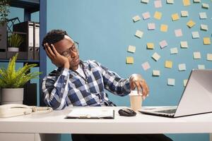 Tired marketing specialist sitting at desk, reviewing key performance indicators on laptop, deep in thought. Exhausted manager resting his head on hand and looking at device screen, online learning. photo
