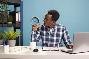 Surprised male manager seated at office desk holding and looking at his clock, running late for meeting. African american businessman with glasses, having a shocked expression after checking the time photo