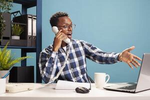 Shocked entrepreneur having serious call with coworker, using his laptop for references. Man with surprised expression is gesturing to device screen as he talks with an employee on his landline phone. photo