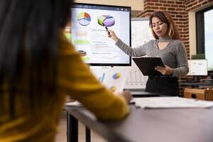 Asian businesswoman reading a document as she points towards the data analytics chart on digital monitor to her colleague. Female manager training her employee, using device screen for presentation. photo