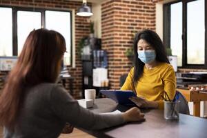 Asian HR manager wearing face mask sits at an office desk, interviewing a job candidate. Female recruiter holds a clipboard, reviewing a CV resume in a startup workspace and questioning an applicant. photo