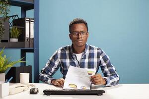 Businessman seated at desk with paperwork, suspicious of the startup project results, having confused expression. POV of male finance analyst checking company accounts on computer, unsure of expenses. photo