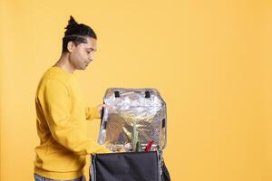 Delivery carrier employee taking takeaway order out of bag, providing it to client. Indian man taking groceries order out of thermal backpack, ordered by customer, studio background photo