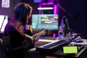 Young singer playing guitar and practicing with online tutorial lessons, looks at daw software on pc. Musician learning new acoustic instrument accords, having phone with greenscreen display. photo