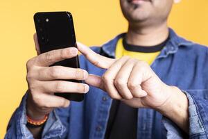 Adult Indian man with a man bun casually using his smartphone, browsing through online apps and social media. Guy with relaxed facial expression shows enjoyment against studio background. Close up. photo