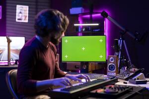 Audio engineer playing electronic piano keyboard in home studio, recording tunes an watches computer with greenscreen after creating a melody on midi controller. Artist uses synthesizer notes. photo