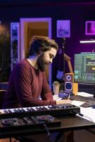Young composer writing rhymes and lyrics to create new song in home studio, thinking about harmonic notes and lines to make cool music. Audio engineer producing soundtracks with gear. photo