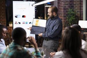 Caucasian male company advisor presents business analytics in laptop during late night group meeting. Diverse colleagues analyzing financial reports and brainstorming strategies in startup office. photo