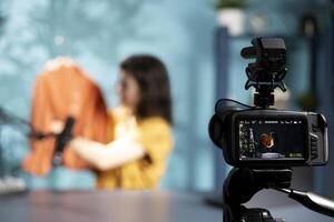 Focus on camera used to film media star in blurry background holding orange jacket in hand, showing fans purchases in studio. Close up of equipment recording content creator presenting new acquisition photo