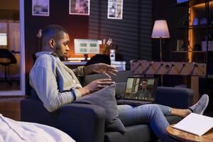 Focused black guy smiling and waving during a web conference in his living room, showcasing online discussion in a digital and wireless lifestyle. Conference speaker with attendees on call. photo