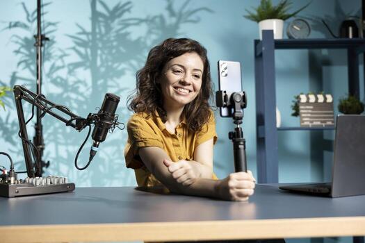 Expressive cheerful content creator using smartphone on selfie stick to shoot . Joyous influencer filming vlog with mobile phone in studio, using professional microphone photo