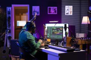 Confident music composer mixing sounds on digital audio software, creating a cool atmosphere in his home studio. Young cheerful producer composing new soundtracks with stereo gear. photo