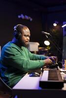 African american music producer twisting knobs on mixing console, producing new tracks with sound effects. Artist composing songs in post production, operates on digital software in home studio. photo