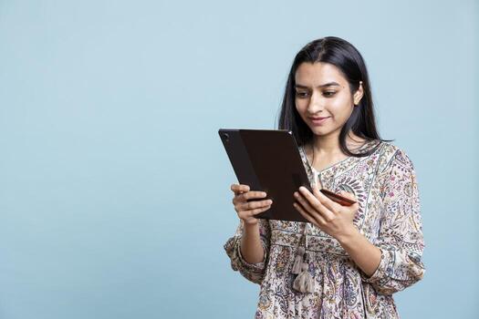 Indian person excited to purchase stuff adding payment method on website with her tablet, isolated over studio background. Ecstatic client does internet shopping, typing debit card info. photo