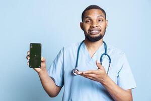 Portrait of joyful african american nurse presenting isolated mockup screen on phone. Smiling healthcare staff pointing at his smartphone with copy space display, studio backdrop. photo