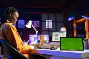 Teleworker in home office checks statistics on laptop, crosschecking with data on green screen tablet. Person using notebook and mockup chroma key device, analyzing company revenue numbers photo