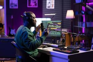 Confident musician singing his own song with a phone to read his verses, using a microphone in home studio to record his track and edit it in post production. Happy artist producer making music. photo