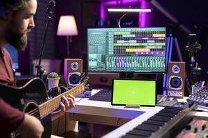 Producer learning to play acoustic guitar with online lessons, playing new accords in his home studio and recording on daw software. Audio engineer singing on on instrument, greenscreen on tablet. photo